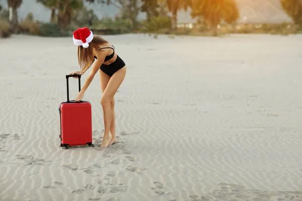 身穿泳衣 头戴圣诞帽的圣诞老人女士 手持红色行李箱 在海滩上挂着一个带着热带棕榈和阳光背景的模型 圣诞节和新年的旅行理念 折扣旅行 — 图库照片