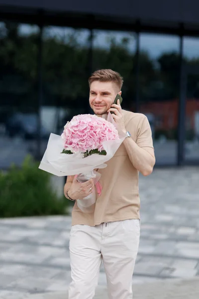 Flower delivery. Smiling delivery man with luxury bouquet of hydrangea flowers talks on smartphone with client on territory modern residential building. Gift for Valentines day, Birthday, Anniversary.