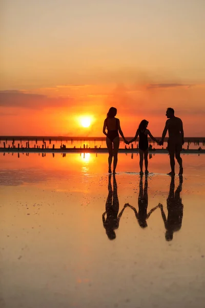 Happy family day. Silhouette of young mom, dad and little daughter holding hands walking together on beach on sunset in summer travel holiday. International Children\'s Day. Concept of family values