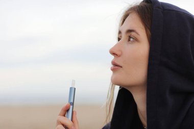Elektronik sigara teknolojisi. Genç bir kadın sigara içiyor ve hibrit sigara cihazından buhar çıkarıyor. Gerçek tütün doldurma yastığı ve tütün ısıtma sistemi kullanıyor. Kötü kötü alışkanlık.