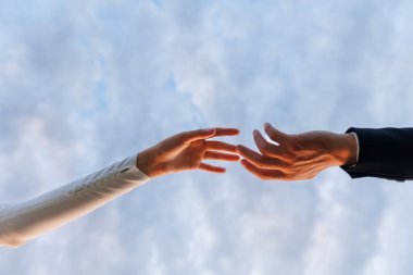 Beyaz elbiseli gelinin elleri ve takım elbiseli damadın elleri birbirine uzanıyor, mavi gökyüzü arka planında parmaklarına dokunuyor. İnsanları kurtarmak ve desteklemek için yardım etmek. Düğün günü. Sevgililer Günü