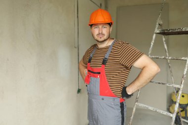 Yenileme dairesi. Kendine güveni tam genç bir ustabaşı tamircinin portresi güvenlik kaskı ve tulumuyla kameraya bakıyor. Boş beton duvarlar, tamir evleri, arka planda merdiven.