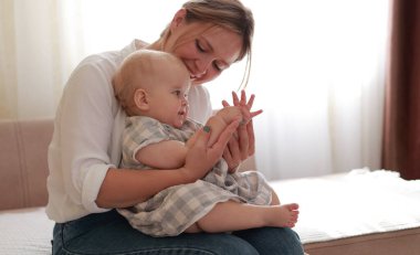 Anneler Günü. Gülümseyen genç anne evde, yatak odasında erkek bebekle el ele oynuyor. Sevgi dolu bir anne, küçük çocuğuyla ilgilenip onu eğitiyor. Sağlıklı çocuk bakımı. Annelik Ebeveynliği