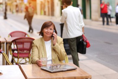 Olgun bir ofis çalışanı, elinde bir fincan kahve ve dizüstü bilgisayarla terasta otururken akıllı telefonuyla yaklaşan iş seyahatinden bahseder.