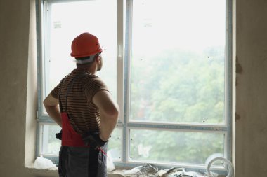 Yenileme dairesi. Yeni binadaki yeni bir daireye yerleştirildikten sonra inşaat işçisinin kasketli arka görüntüsü plastik pencereye bakıyor ve kontrol ediyor. İnşaat alanı, endüstriyel iş.