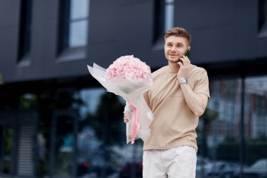 Çiçek teslimatı. Elinde bir buket ortanca çiçeği olan gülümseyen teslimatçı müşterisiyle modern apartmanda akıllı telefondan konuşuyor. Sevgililer günü, doğum günü, yıldönümü hediyesi..