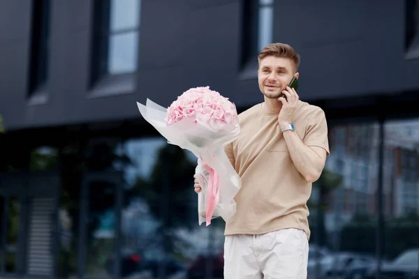 Flower delivery. Smiling delivery man with luxury bouquet of hydrangea flowers talks on smartphone with client on territory modern residential building. Gift for Valentines day, Birthday, Anniversary.