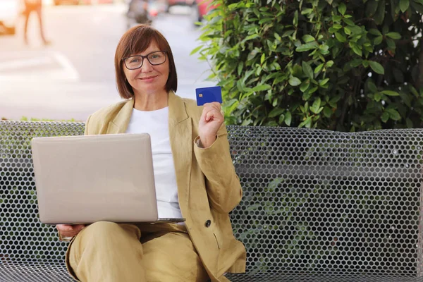 Happy stylish middle aged businesswoman shows credit debit card while e-banking on laptop on bench outdoors. Online payment, shopping online, e-commerce, internet banking, spending money concept.