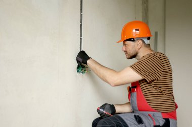 Üniformalı elektrikçi erkek, koruyucu eldivenler ve kask prizdeki elektrik voltajını kontrol ediyor elektrikli tornavida kullanıyor. Kapalı alandaki voltaj detektörüyle prizdeki kabloları inceliyor..