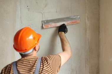 Rear view of handyman worker in overalls and protective helmet plastering concrete wall with putty using a big putty knife. Renovation at apartment and construction new building concept clipart