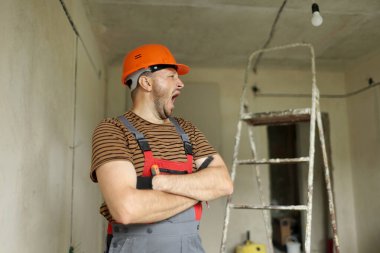 Yenileme dairesi. Koruyucu kasklı ve tulumlu çapraz elli genç ustabaşı tamirci esniyor. Boş beton duvarlar, tamir evleri, arka planda merdiven.