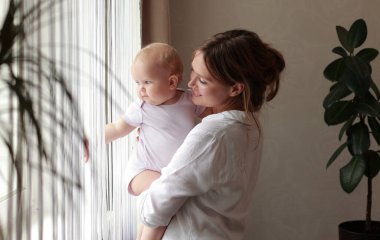 Sevgi dolu genç bir anne kucağında sevimli bir kız bebek kucağında küçük bir çocuğa sarılıyor. Mutlu şefkatli bir anne, evde pencerenin önünde duran küçük çocuğa sarılıyor. Mutlu annelik kavramı.