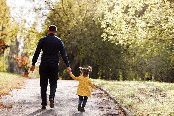 Happy fathers day. Rear view of dad holds hand his little daughter and walks outdoors at autumn park. Single daddy with child relaxes and spends time together. Family day and childhood concept