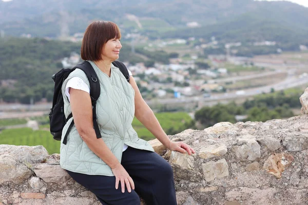 Active middle aged woman with backpack relaxes sits on hill, looks at beautiful view after hiking up. Healthy lifestyle in retirement. Adventure seeks woman treks enjoys freedom.