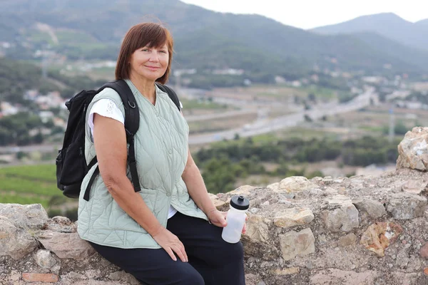 Adult active woman travel with backpack relaxes sit with water bottle on hill after hiking up, look at camera. Healthy lifestyle in retirement. Adventure seeks woman treks enjoys freedom and vacation.
