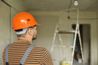 Güvenlik kaskı ve tulumlu profesyonel tamirci müteahhidi boş beton duvarlara bakıyor ve yenileme odasını planlıyorlar. Tamir evi kavramı. Arka planda merdiven.