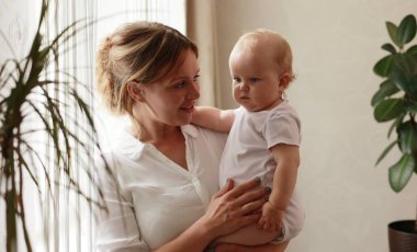 Sevgi dolu genç bir anne kucağında sevimli bir kız bebek kucağında küçük bir çocuğa sarılıyor. Mutlu şefkatli bir anne, evde pencerenin önünde duran küçük çocuğa sarılıyor. Mutlu annelik kavramı.