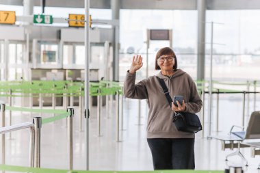 Happy smiling mature traveler woman hold smartphone and wave hand in greeting someone who meeting her at airport terminal after arrived from trip at new country at holiday vacation. Retirement travel. clipart