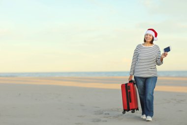 Noel Baba şapkalı mutlu orta yaşlı kadın kırmızı valizli, pasaport kartıyla sahilde deniz kenarında. Mutlu yıllar 2023 bayram konsepti. Kış tatili.