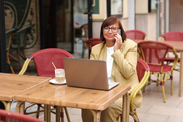 Olgun bir ofis çalışanı, elinde bir fincan kahve ve dizüstü bilgisayarla terasta otururken akıllı telefonuyla yaklaşan iş seyahatinden bahseder.