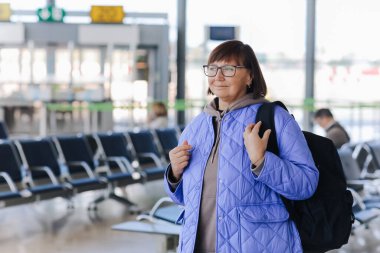 Orta yaşlı gözlüklü, el çantalı, sırt çantalı bir kadın havaalanının kapısında elinde akıllı bir telefonla bekliyor. Aktif macera, hayat tarzı seyahatini emekliye ayırdı