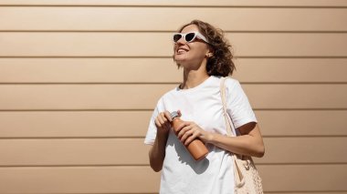 Neşeli bin yıllık genç kadın yeniden kullanılabilir metal bir şişe açıyor ve güneşli yaz gününde açık havada modern bina arka planında temiz su içiyor. Sıfır atık, çevre dostu, plastik kavram yok..