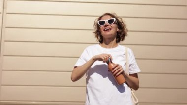 Neşeli bin yıllık genç kadın yeniden kullanılabilir metal bir şişe açıyor ve güneşli yaz gününde açık havada modern bina arka planında temiz su içiyor. Sıfır atık, çevre dostu, plastik kavram yok..