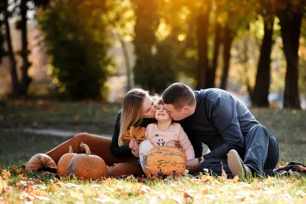 Family day. Happy mom and dad kissing little child daughter, having fun sit on grass with pumpkins at autumn park, enjoying spend time together at weekend. Parental care and happy carefree childhood.