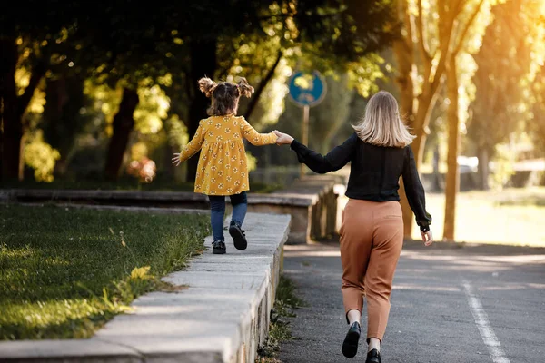 Mothers day. Happy young mom is holding by hand and walking with adorable little daughter at autumn park. Mom with kid are having fun, playing at weekend. Outdoors vacation activity. Children's day.