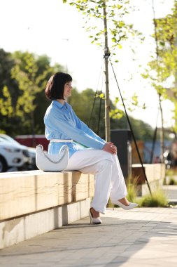 Kısa saçlı, zarif, gülümseyen esmer kadın mavi gömlek, beyaz pantolon ve şık bir çanta giyer modern şehir caddesinde ahşap bankta oturur ve güneşli sıcak bir güne bakar. İş resmi biçimi