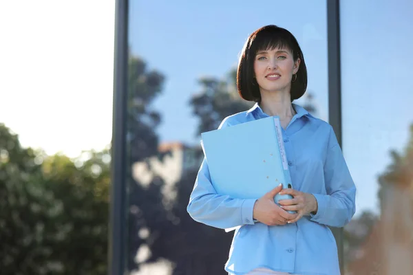 Business school concept. Smiling confident business woman student in blue shirt holds document files folder, smiles and looks at camera while standing outdoors by the modern building company