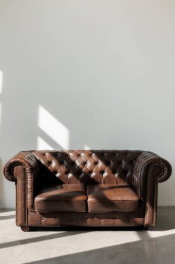 Vintage quilted classic brown sofa against white wall with sunlights from window on concrete floor at studio. Natural leather with rhombic stitching with buttons. Furniture design. Copy space. clipart