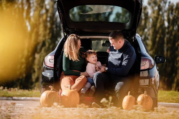 Family day. Happy mom, dad and little child daughter sit at car trunk, look at camera at autumn park with pumpkins, enjoy spend time together at weekend. Parental care and happy carefree childhood.