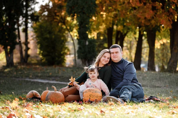 Thanksgiving day. Happy mom, dad and little child daughter having fun sit on grass with pumpkins at autumn park, enjoying spend time together at weekend. Parental care and happy carefree childhood.