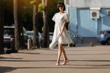 Zarif, genç, esmer, yaz tarzı kıyafetler giymiş, gülümseyen, şehirde mutlu, güneş gözlüğü takan, kısa beyaz elbiseli, çanta ve yüksek topuklu ayakkabı giyen bir kadın. Dünya Kadınlar Günü. Romantik ruh hali.