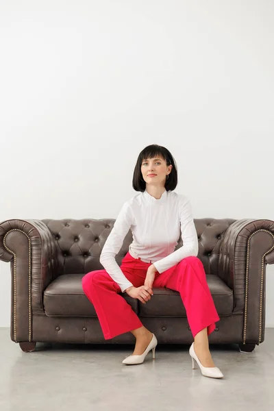 Positive brunette middle aged woman wears elegant red pants, white blouse and high heels confidently looks at camera, sits relaxed on classic leather sofa at empty studio room. World women\'s day.