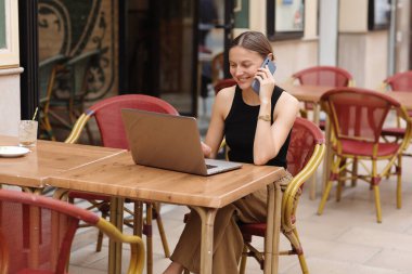 Açık kafedeki laptopta çalışırken akıllı telefonda başarılı bir şekilde gülümseyen genç iş kadını iletişimi. Kamu alanından uzak çalışma kavramı, dijital serbest çalışma ve modern yaşam tarzı.