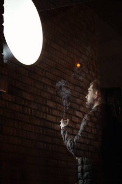 Sakallı bir adam, alacakaranlıkta ağır iş gününden sonra dışarıda sigara içmekten zevk alıyor. Cam kapının arkası. Sigara can sıkıntısından öfke kontrolüne kadar her şeye iyi gelir.