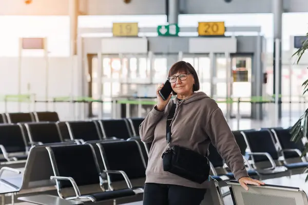 Happy smiling mature traveler woman hold smartphone and wave hand in greeting someone who meeting her at airport terminal after arrived from trip at new country at holiday vacation. Retirement travel.