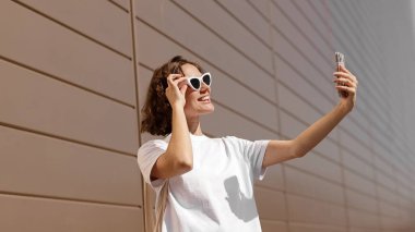 Güneş gözlüklü mutlu genç kadın güneşli bir yaz gününde akıllı telefondan selfie çekiyor. Kadın blogcu cep telefonu kamerası kullanıyor. Çekici beyaz kız saçına dokunup selfie çekiyor.