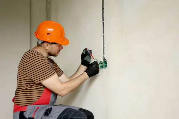 Electrician male in uniform, protective gloves and helmet checks presence of electrical voltage in socket phase uses electrical tester screwdriver.Examining wires in outlet by voltage detector indoor.
