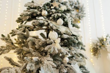 Yapay karlı bir Noel ağacı, şenlikli dekorasyonlar, parıltılı beyaz ksenler, parlak mücevherler, toplar, parlak çiçek süslemeleri. Altın ışıklı çelenkler arka planda yanar. Yeni yıl 2025 bayramı