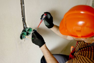 Üniformalı elektrikçi erkek, koruyucu eldivenler ve kask prizdeki elektrik voltajını kontrol ediyor elektrikli tornavida kullanıyor. Kapalı alandaki voltaj detektörüyle prizdeki kabloları inceliyor..