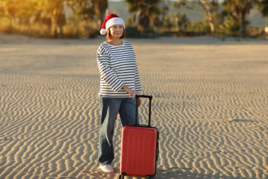 Noel Baba şapkalı, çizgili tişörtlü, 50-55 yaş arası mutlu bir yetişkin kadın kum sahili kenarında kırmızı bavul tutuyor. Palmiyeli yeni yıl tatilini tropikal tatil beldesinde yalnız kutluyor..