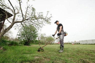Koruyucu giysili profesyonel bahçıvanın yandan görünüşü yeşil çimenleri buduyor. Erkek işçi, el elektriği veya benzin çim biçme makinesi ile yabani otları biçer, şehir parkında ya da arka bahçede çim biçer. Bahçe bakımı araçları.