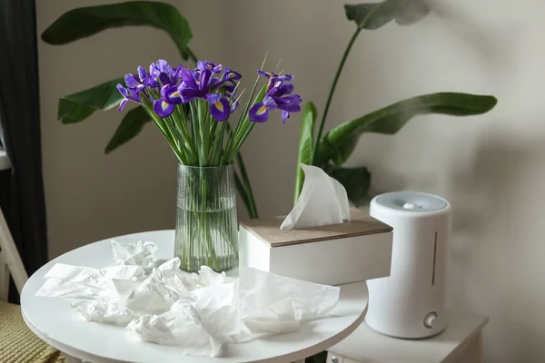 Seasonal allergy rhinitis to flowers. Bouquet of purple iris flowers in vase on table, many used napkins and humidifier or air purifier at home interior. Allergic symptom of odor pollen flowering.