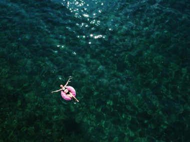 Açık kollu, mayo giymiş, yüzen ve dinlenen genç bir kadının havadan drone görüntüsü. Su yüzeyinde şeffaf bir tabanla yüzüyor. Egzotik tatil beldesinde yaz tatili.