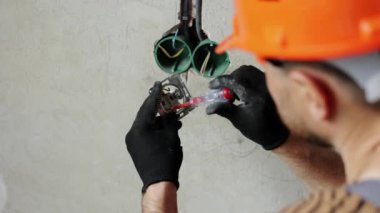 Üniformalı elektrikçi erkek, koruyucu eldivenler ve kask prizdeki elektrik voltajını kontrol ediyor elektrikli tornavida kullanıyor. Kapalı alandaki voltaj detektörüyle prizdeki kabloları inceliyor..
