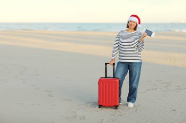 Noel Baba şapkalı gülümseyen olgun kadın sahilde pasaport, bilet ve kırmızı bavul taşıyor. Mutlu yıllar 2024 bayram kutlamaları. Kış tatili