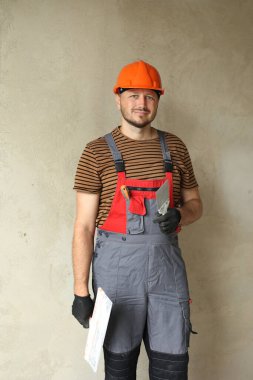 Tulumlu ve koruyucu turuncu kasklı gülümseyen tamirci macunla beton duvarı sıvadığı için iki spatula tutuyor. Apartman onarımı ve yeni bina konsepti. Boşluğu kopyala.
